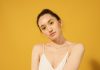 Close-up portrait of a woman with yellow makeup, hoop earrings, and spaghetti strap top against a yellow background.