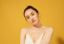 2026 Yaz Sezonunun Öne Çıkan 5 Makyaj Trendi Close-up portrait of a woman with yellow makeup, hoop earrings, and spaghetti strap top against a yellow background.