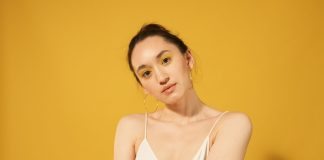 Close-up portrait of a woman with yellow makeup, hoop earrings, and spaghetti strap top against a yellow background.