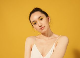 Close-up portrait of a woman with yellow makeup, hoop earrings, and spaghetti strap top against a yellow background.