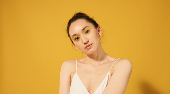 2026 Yaz Sezonunun Öne Çıkan 5 Makyaj Trendi Close-up portrait of a woman with yellow makeup, hoop earrings, and spaghetti strap top against a yellow background.