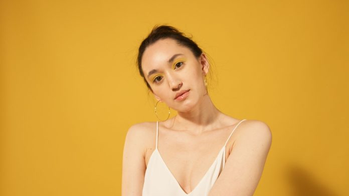 2026-yaz-sezonunun-one-cikan-5-makyaj-trendi-1 Close-up portrait of a woman with yellow makeup, hoop earrings, and spaghetti strap top against a yellow background.