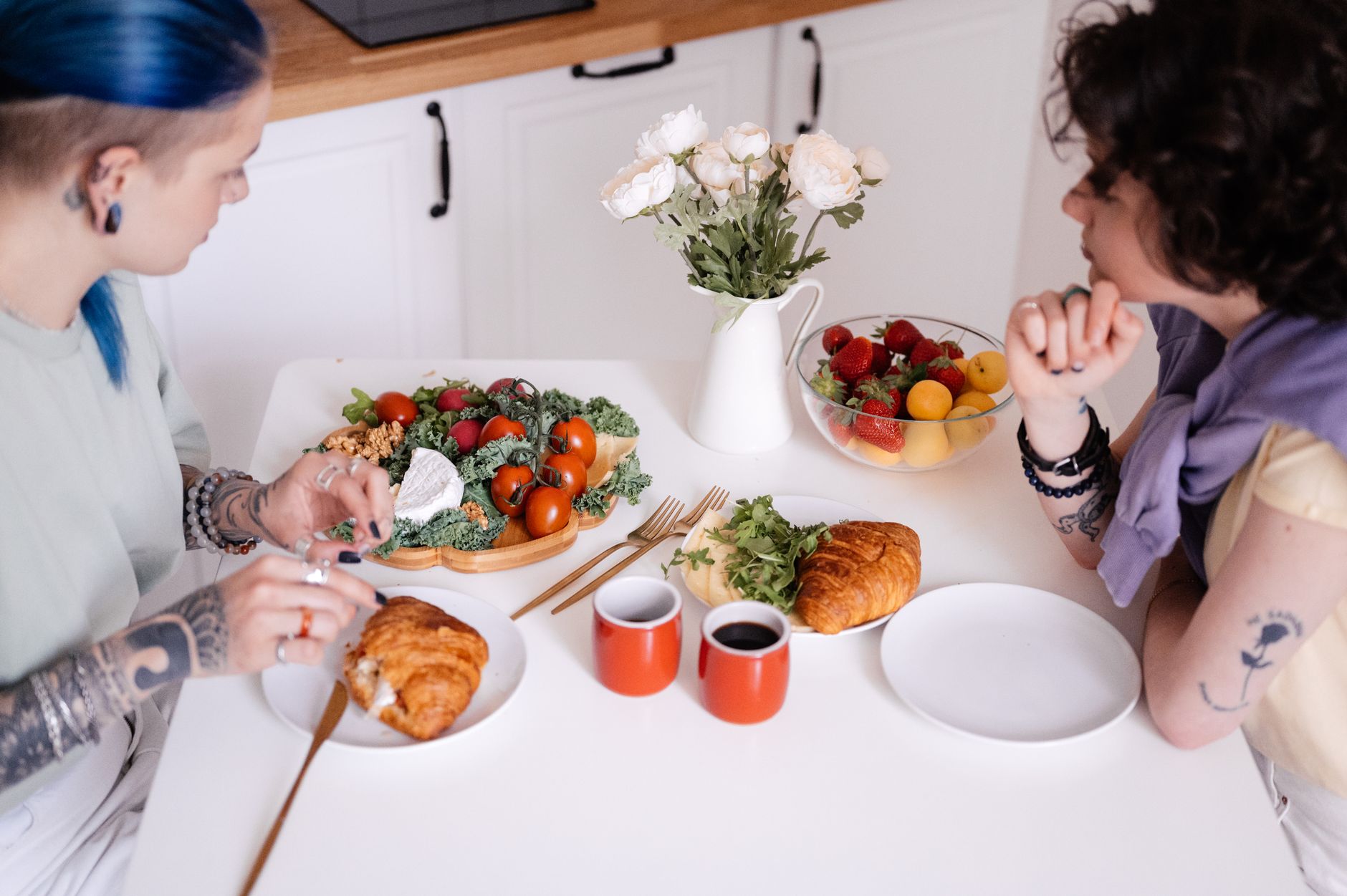 Two women share a healthy breakfast, featuring fresh fruit, coffee, and croissants, on a kitchen table.