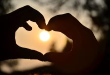 Yaz Ortası Aşk Rüzgarları: Kalbinizin Kapısını Kim Çalacak? Silhouette of hands forming a heart shape around a glowing sunset outdoors.
