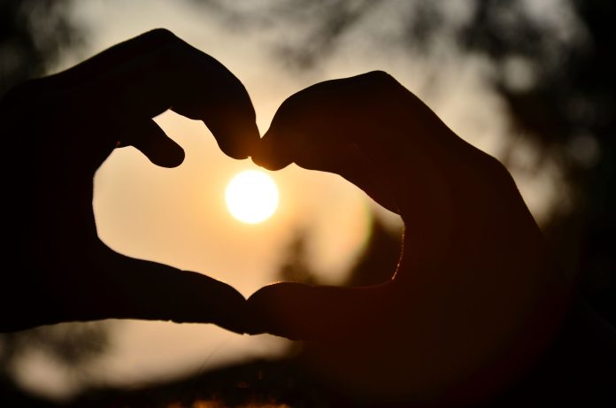 Silhouette of hands forming a heart shape around a glowing sunset outdoors.